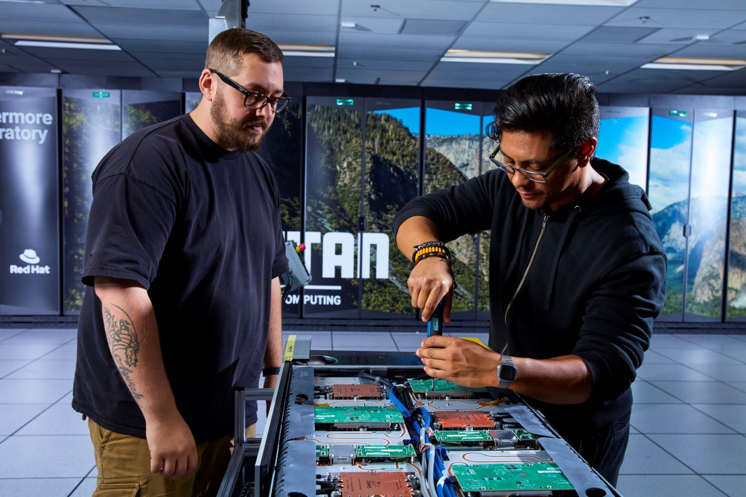 Two men stand over a server board in front of the El Capitan supercomputer.