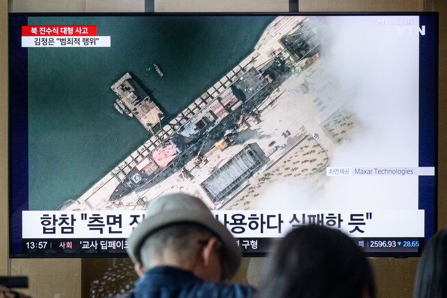 In a photo taken in Seoul on May 22, 2025, people sit near a television showing news footage with a satellite still image courtesy of Maxar Technologies showing a general view of a new North Korean warship at the harbour ahead of its launch in Chongjin.